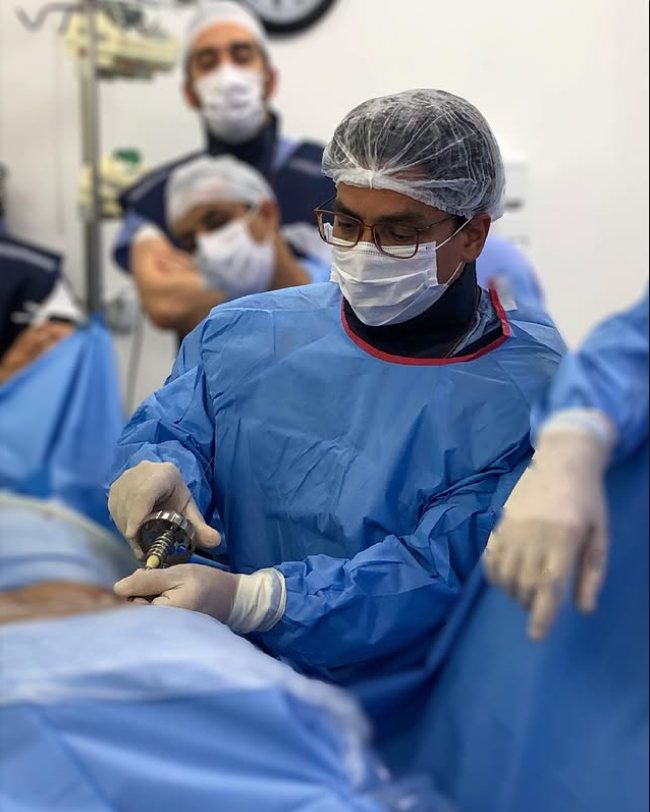 Dr. Márcio Crisanto no centro cirúrgico, de touca, máscara e avental, com a equipe ao fundo